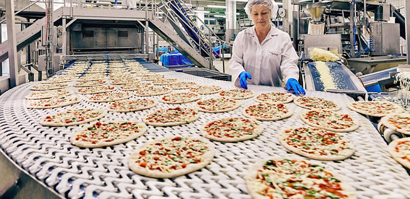 Mitarbeiterin, in Arbeitskleidung, am Fließband mit vielen aufgereihten Pizzen. 
