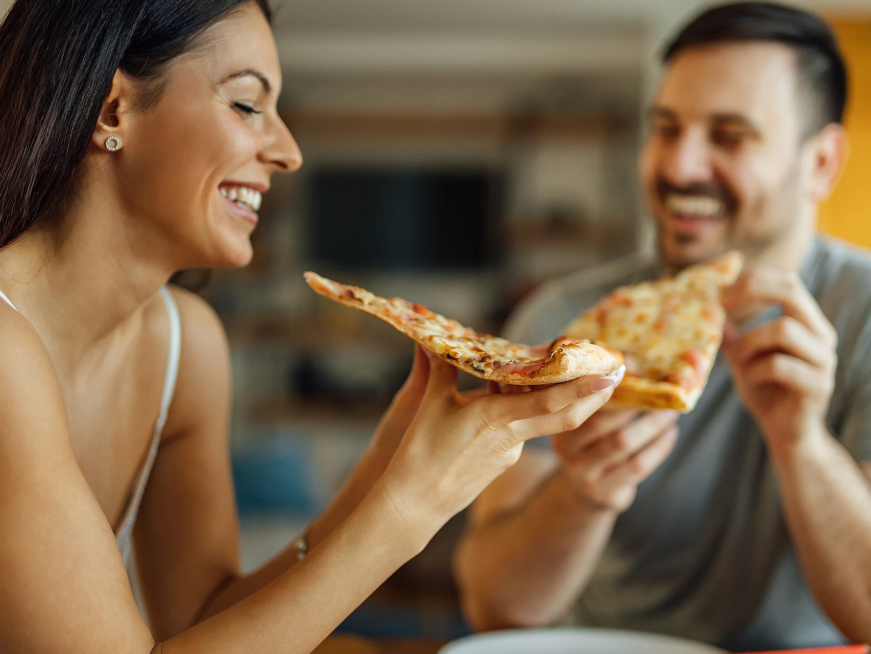 Eine Frau im Fokus und ein Mann im Hintergrund sitzen am Tisch und sind beide kurz vorm Abbeißen jeweils eines Pizzastücks.