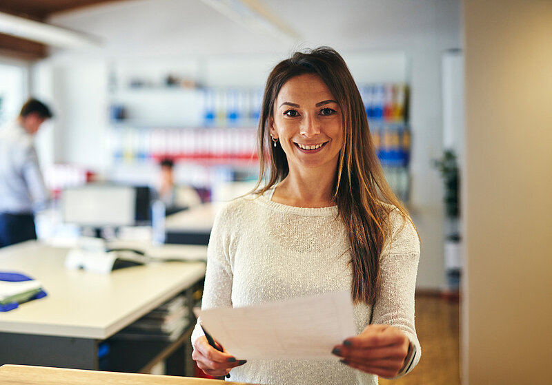 Eine Mitarbeiterin im Büro mit einem Zettel in der Hand, lächelt in die Kamera. 