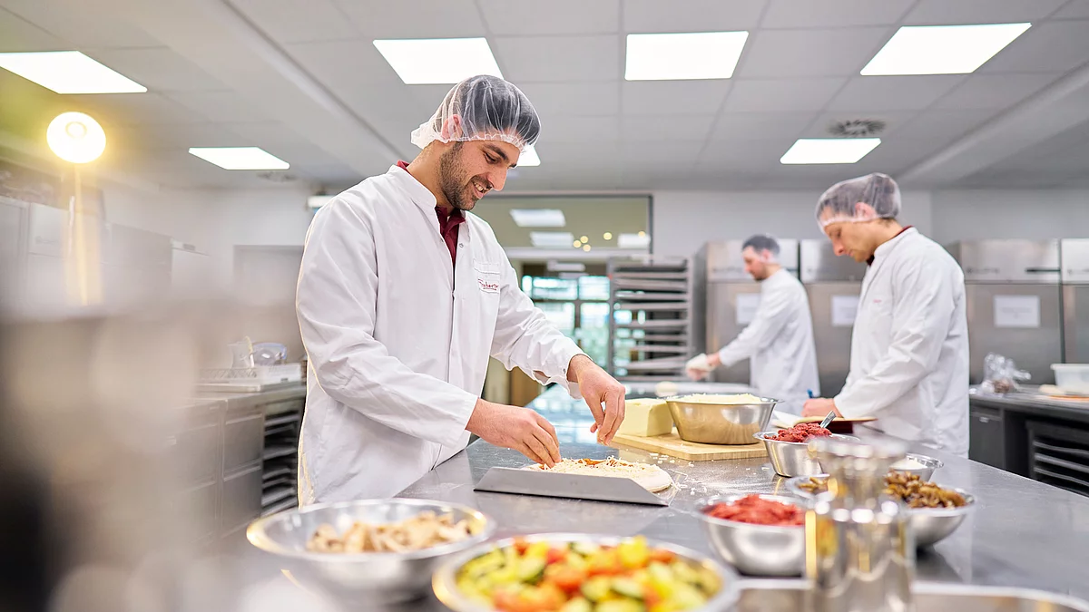 Mitarbeitende in Arbeitskleidung in der Küche mit Pizzen und geschnittenen Lebensmitteln vor sich. 