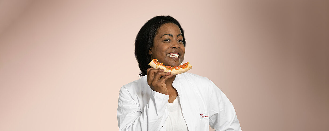 Eine Mitarbeiterin steht lächelnd vor einem schlichten Hintergrund mit einer abgebissenen Pizza.