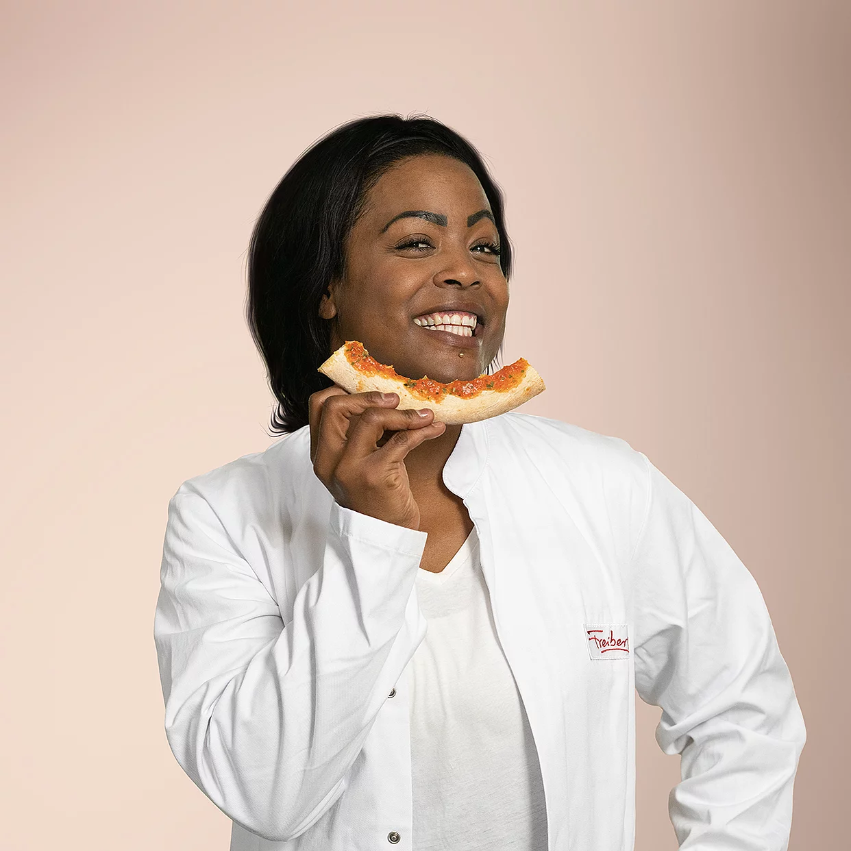 Eine Mitarbeiterin steht lächelnd vor einem schlichten Hintergrund mit einer abgebissenen Pizza.