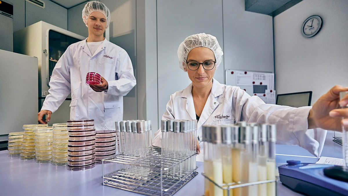 Zwei Mitarbeitende in Arbeitskleidung im Labor vor einem mit Proben befüllten Tisch.