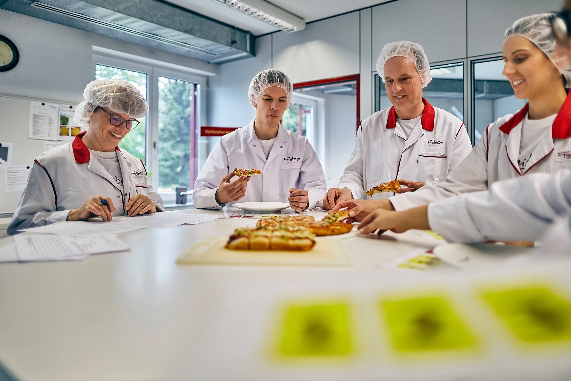 Fünf Mitarbeitende in Arbeitskleidung stehen an einem Tisch und probieren Pizza. Eine Mitarbeiterin schreibt alles mit.