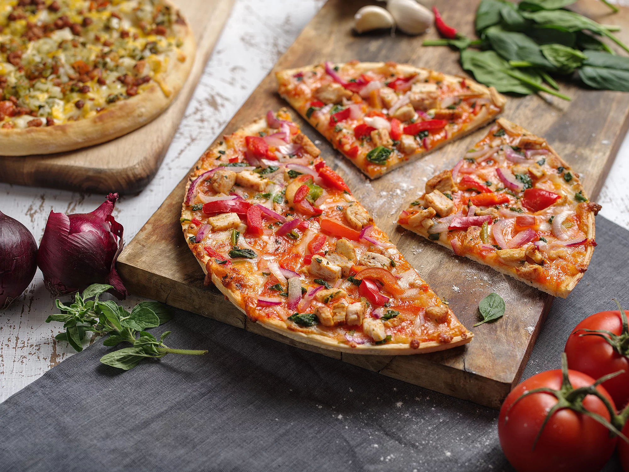 Zwei Pizzen jeweils auf einem Holzbrett. Drumherum dekoriert mit Tomaten, Zwiebeln, Basilikum und Knoblauchzehen.