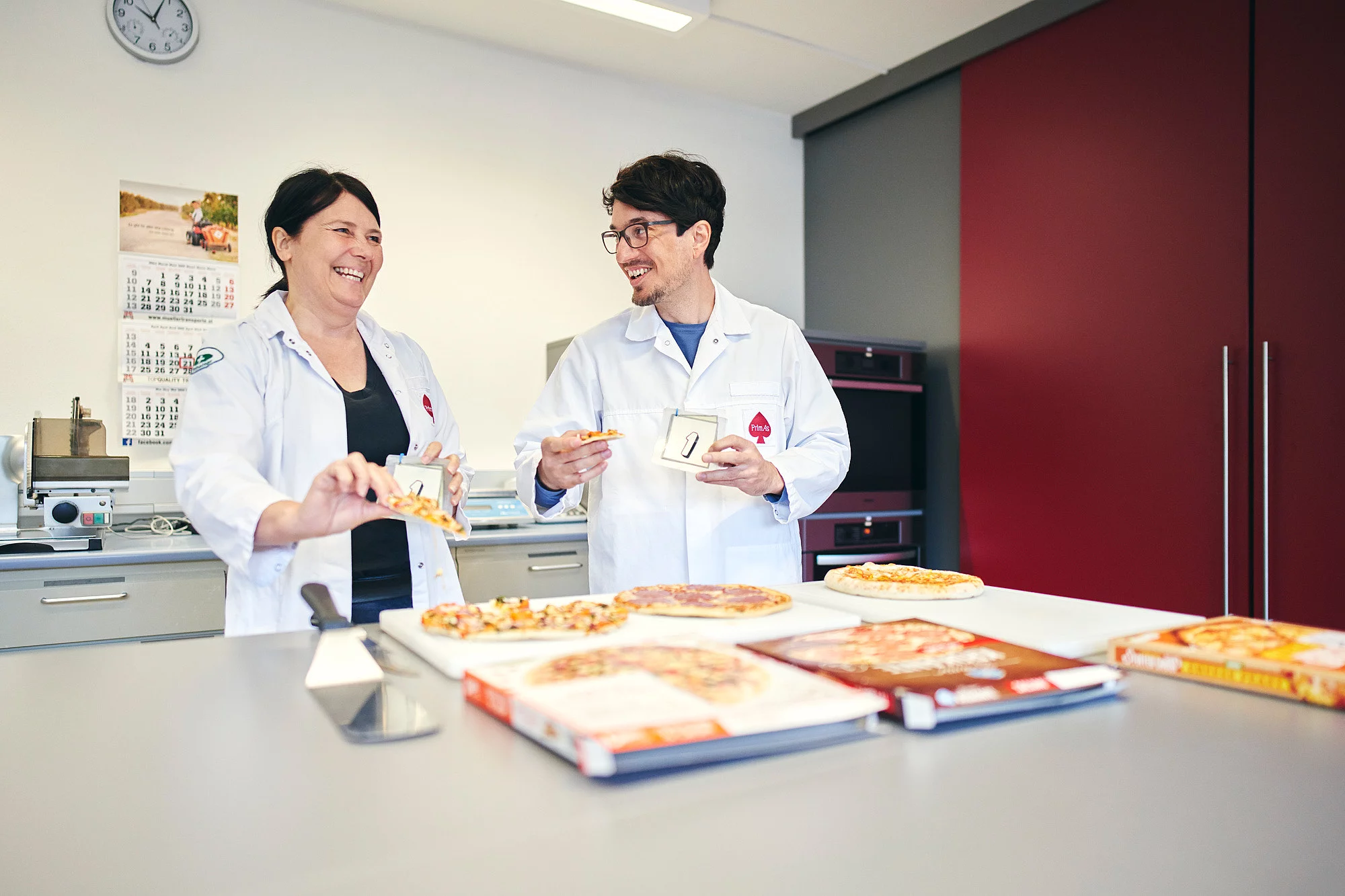 Zwei Mitarbeitende stehen lächelnd an einem Tisch voller Pizza, mit einem Pizzastück in der Hand und einer Karte mit einer 1 darauf abgebildet.