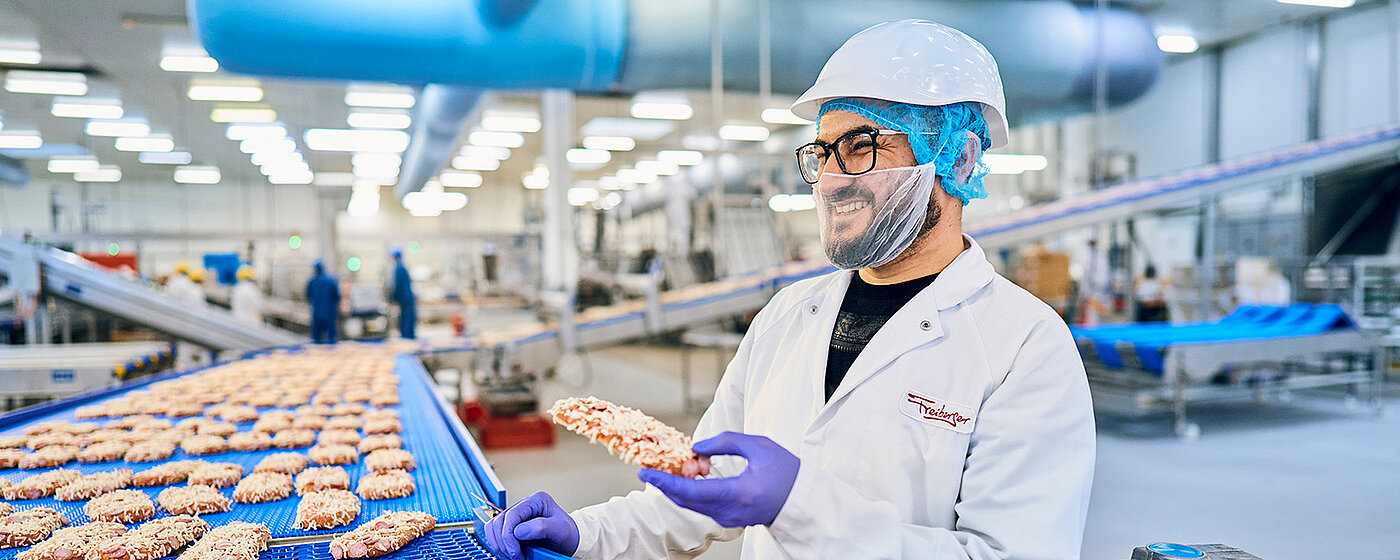 Ein Mitarbeiter am Band in der Produktion mit einem Pizza-Baguette in der Hand 