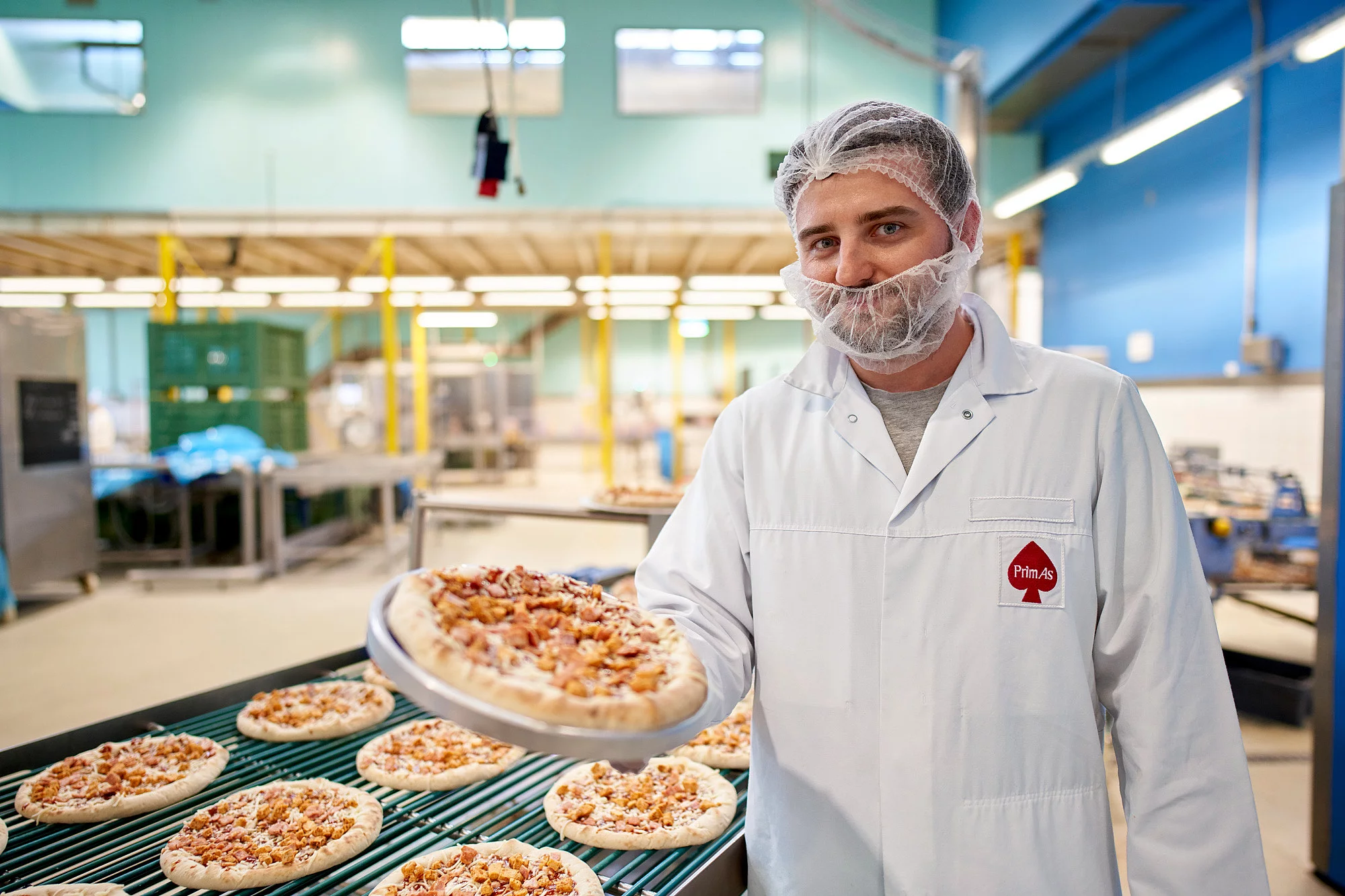 Ein Mitarbeiter in Arbeitskleidung steht vor einer Maschine für die Pizzaproduktion und hält eine Pizza in der Hand.