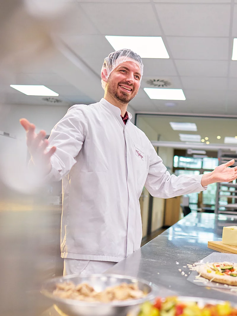 Drei Mitarbeitende stehen in der Industrieküche um einen Tisch und belegen Pizza.