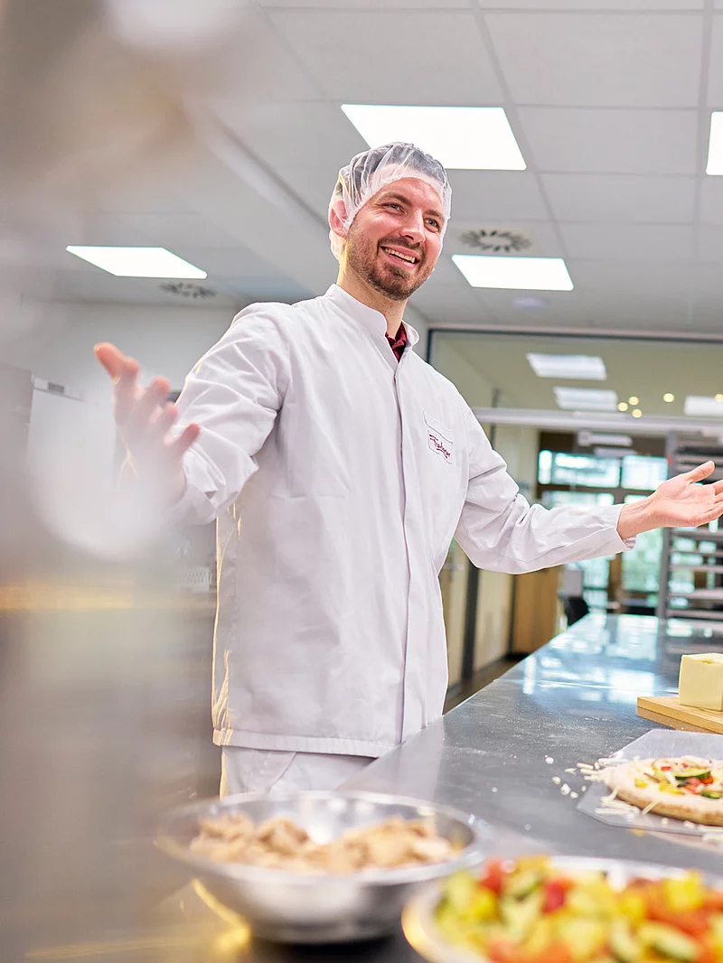Drei Mitarbeitende stehen in der Industrieküche um einen Tisch und belegen Pizza.