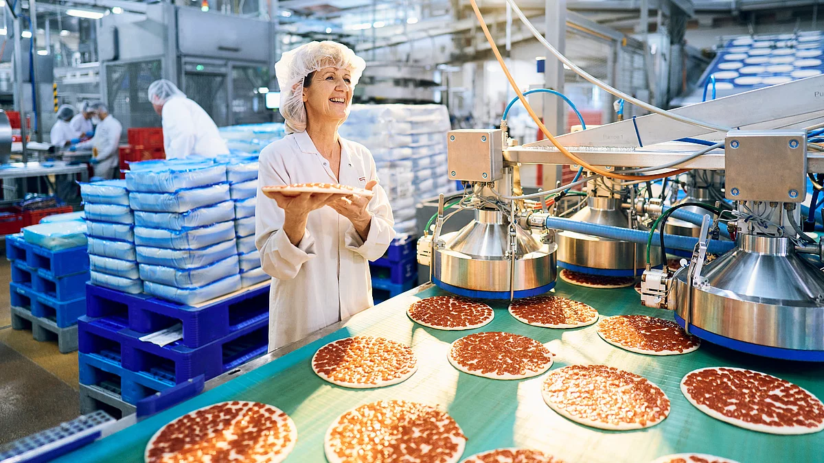Mitarbeiterin in Arbeitskleidung mit Pizzaboden in der Hand am pizzabefüllten Band.