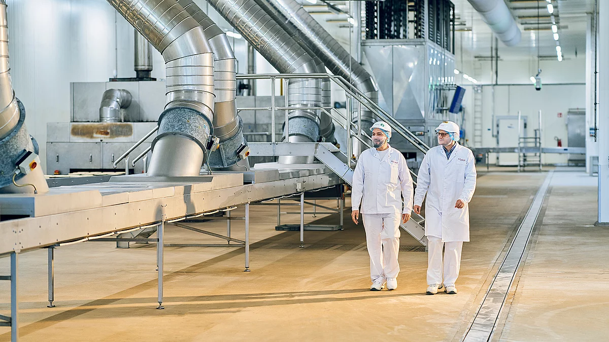 Zwei Mitarbeitende laufen in Arbeitskleidung durch die Produktion.