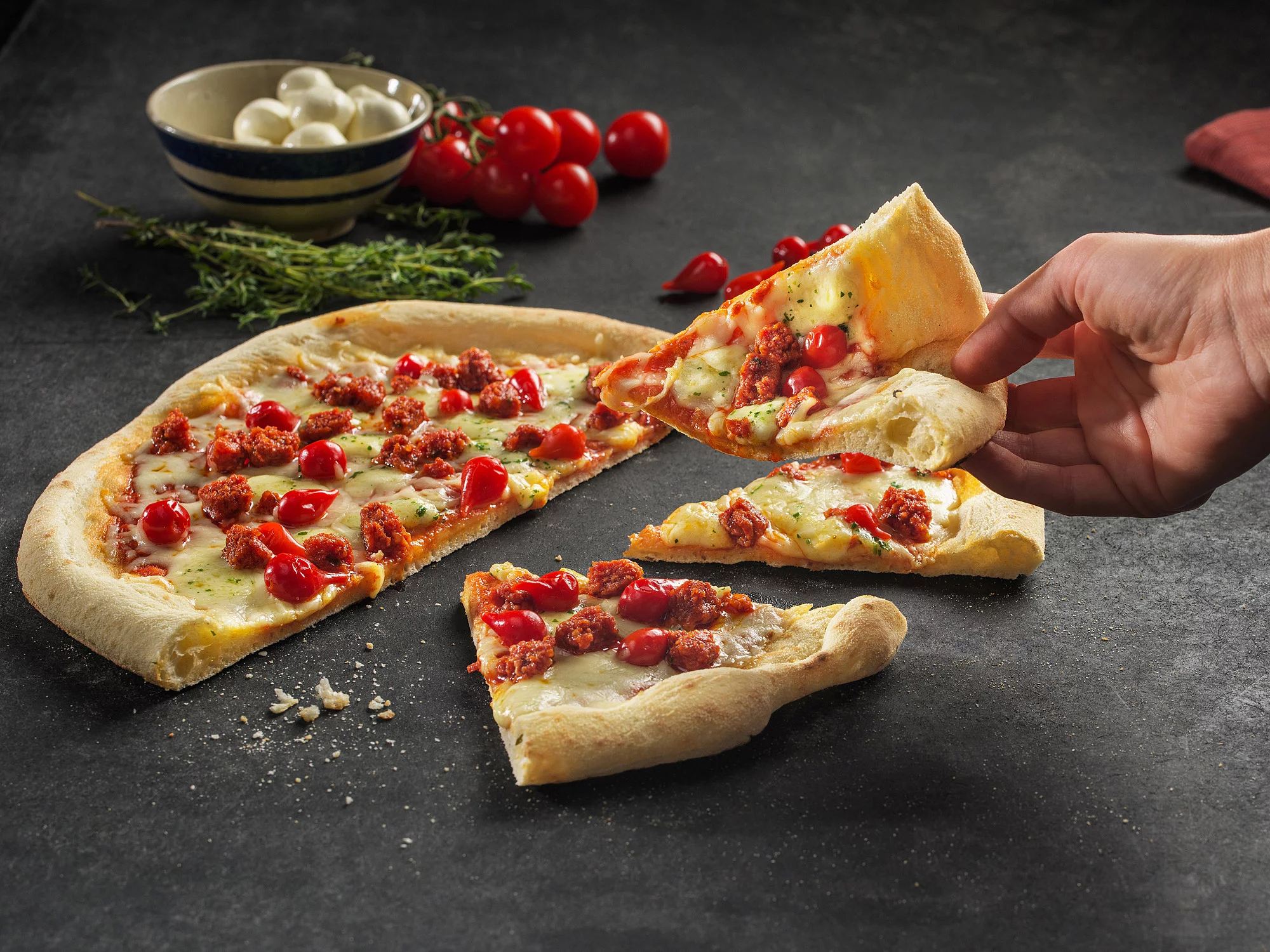 Rechte Hand mit einem Pizzastück in der Hand vor einer Arbeitsplatte mit einer angeschnittenen Pizza.