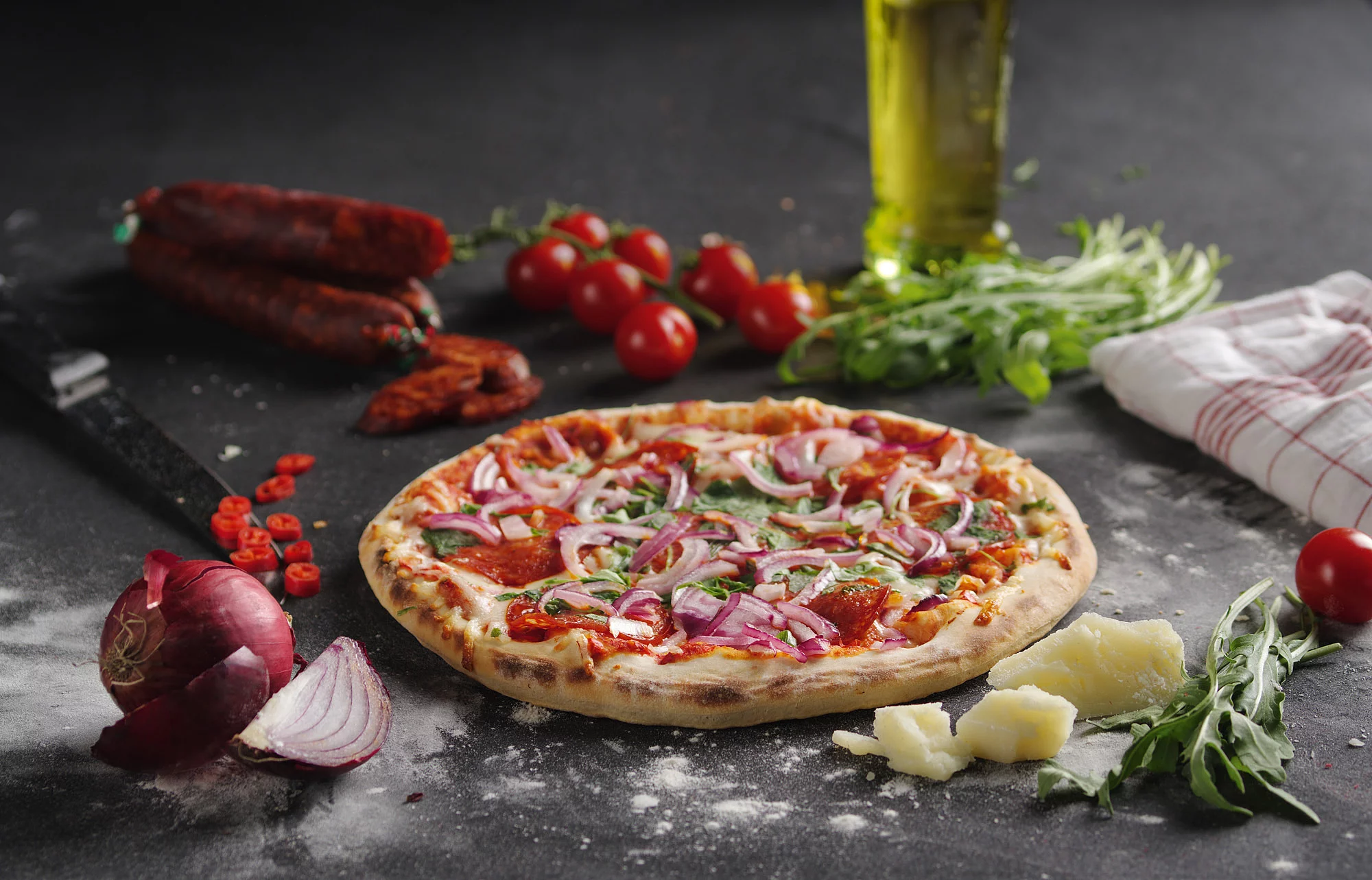 Pizza auf einer Arbeitsplatte drumherum dekoriert mit Tomaten, Parmesan, Salami, Zwiebeln, Basilikum und Olivenöl.