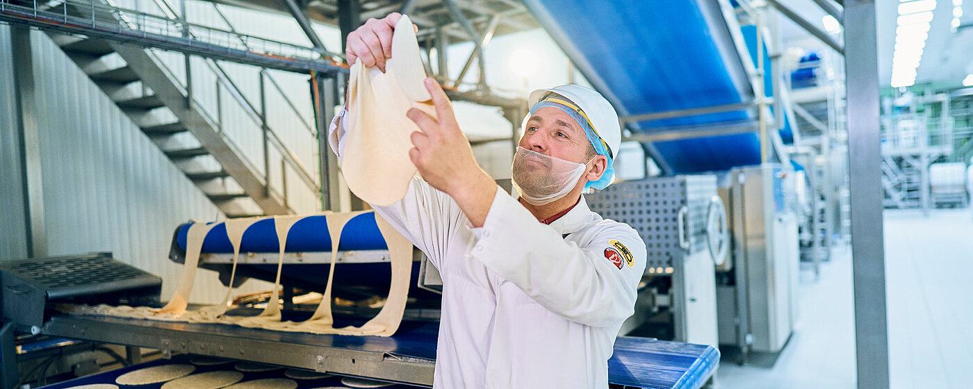 Mitarbeiter in Arbeitskleidung am Produktionsband mit einem Pizzateig in der Hand.