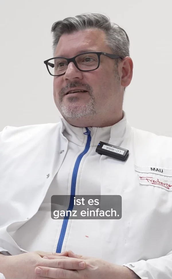 Eine sitzende Person mit weißem Kittel und verschränkten Händen.