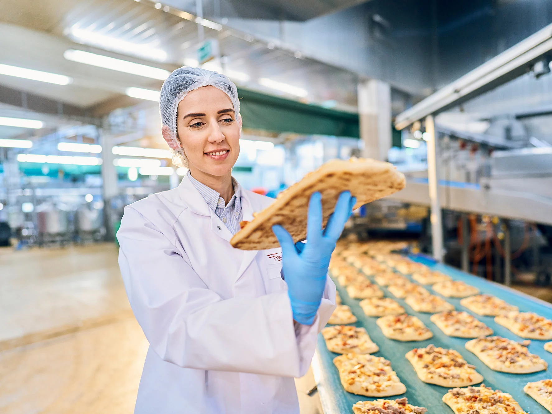 Mitarbeiterin in Arbeitskleidung vor dem Backshop-Pizza-Fließband, mit einem Pizza-Brot in der Hand.