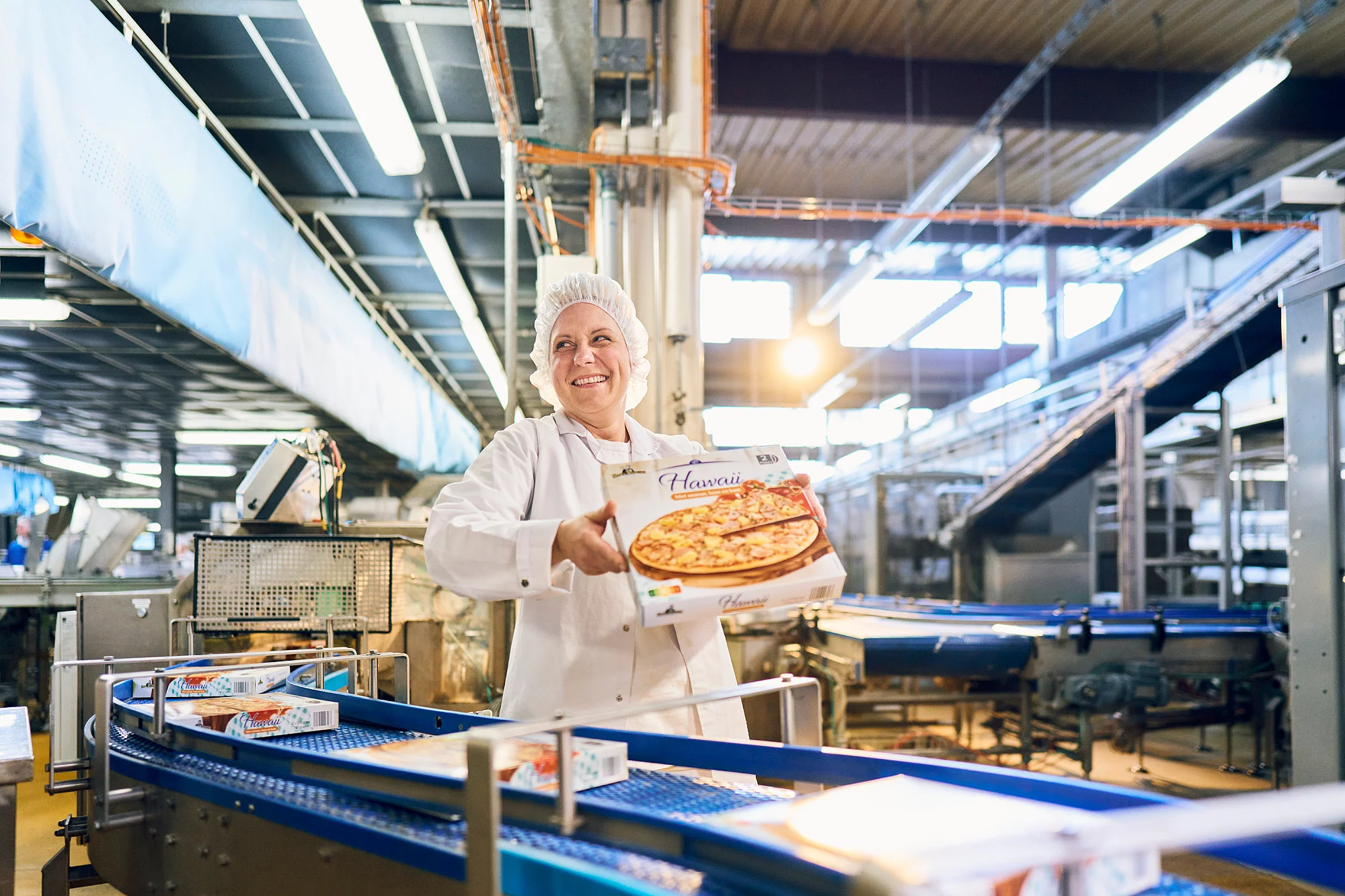 Mitarbeiterin in Arbeitskleidung steht vor einem Fließband und hält einen Pizzakarton hoch.