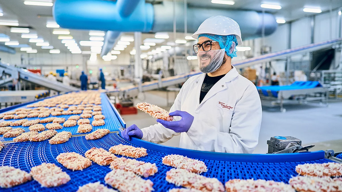 Mitarbeiter in Arbeitskleidung hält ein Pizza-Baguette an dem Produktionsband in der Hand.