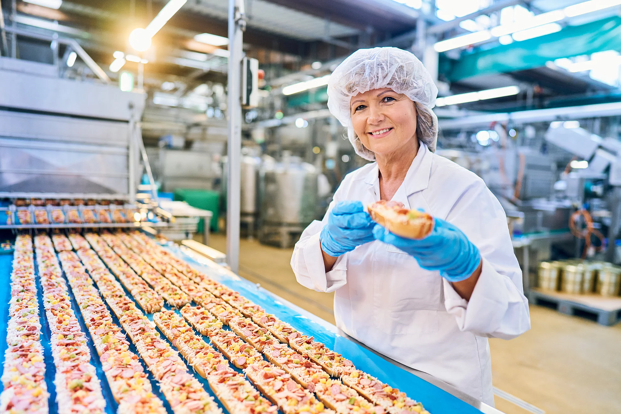Mitarbeiterin in Arbeitskleidung steht vor einem Fließband und hält ein Pizza-Baguette in der Hand.