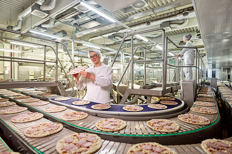 Mitarbeiter in Arbeitskleidung am pizzagefüllten Band in der Betreibshalle.
