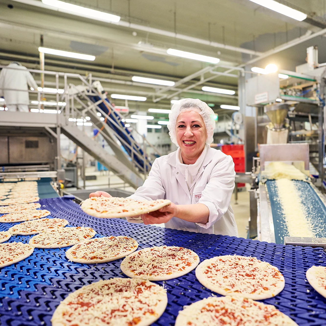 Mitarbeiterin in Arbeitskleidung hält Pizza lächelnd in die Kamera an einem Produktionsband.