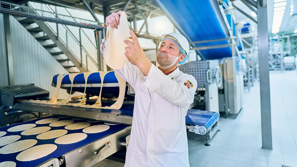 Mitarbeiter in Arbeitskleidung in der Produktion mit einem Pizzateig in der Hand am pizzabefüllten Band.