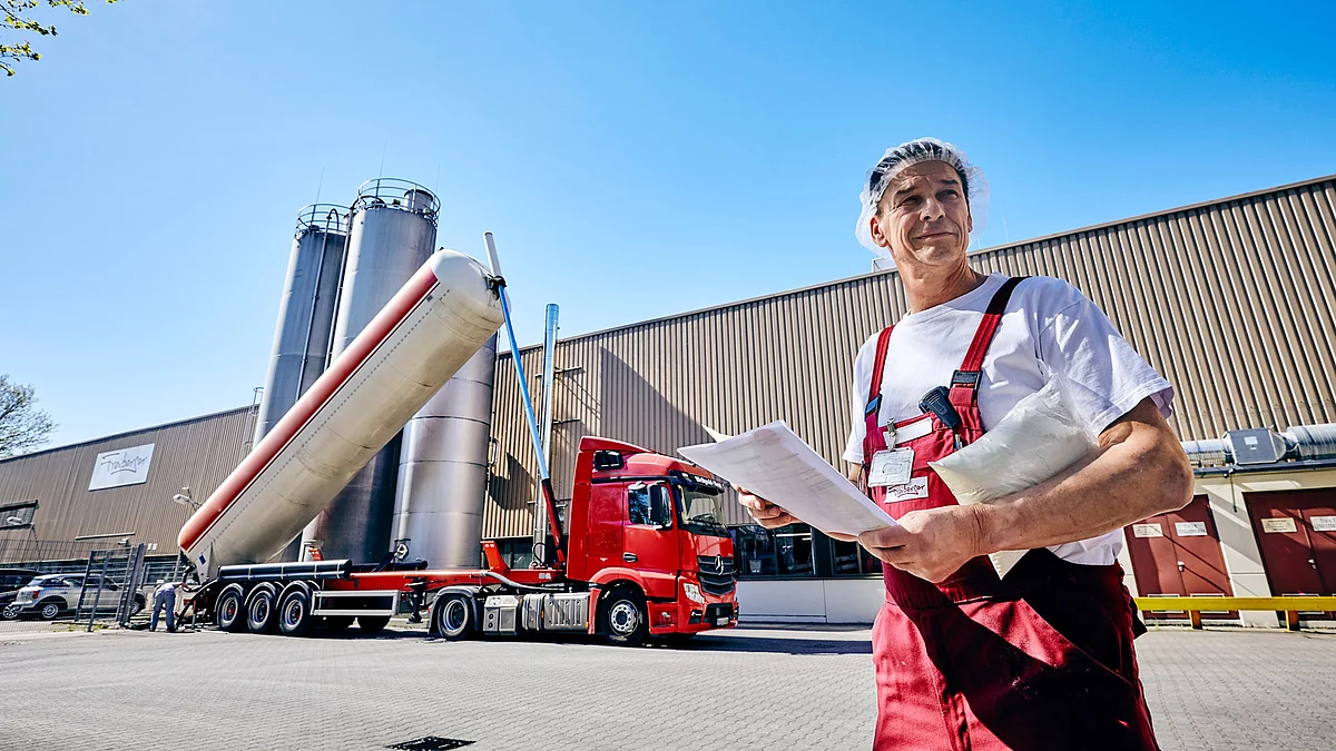 Mitarbeiter in Latzhose mit Plan in der Hand an einer Produktionsstätte draußen. 