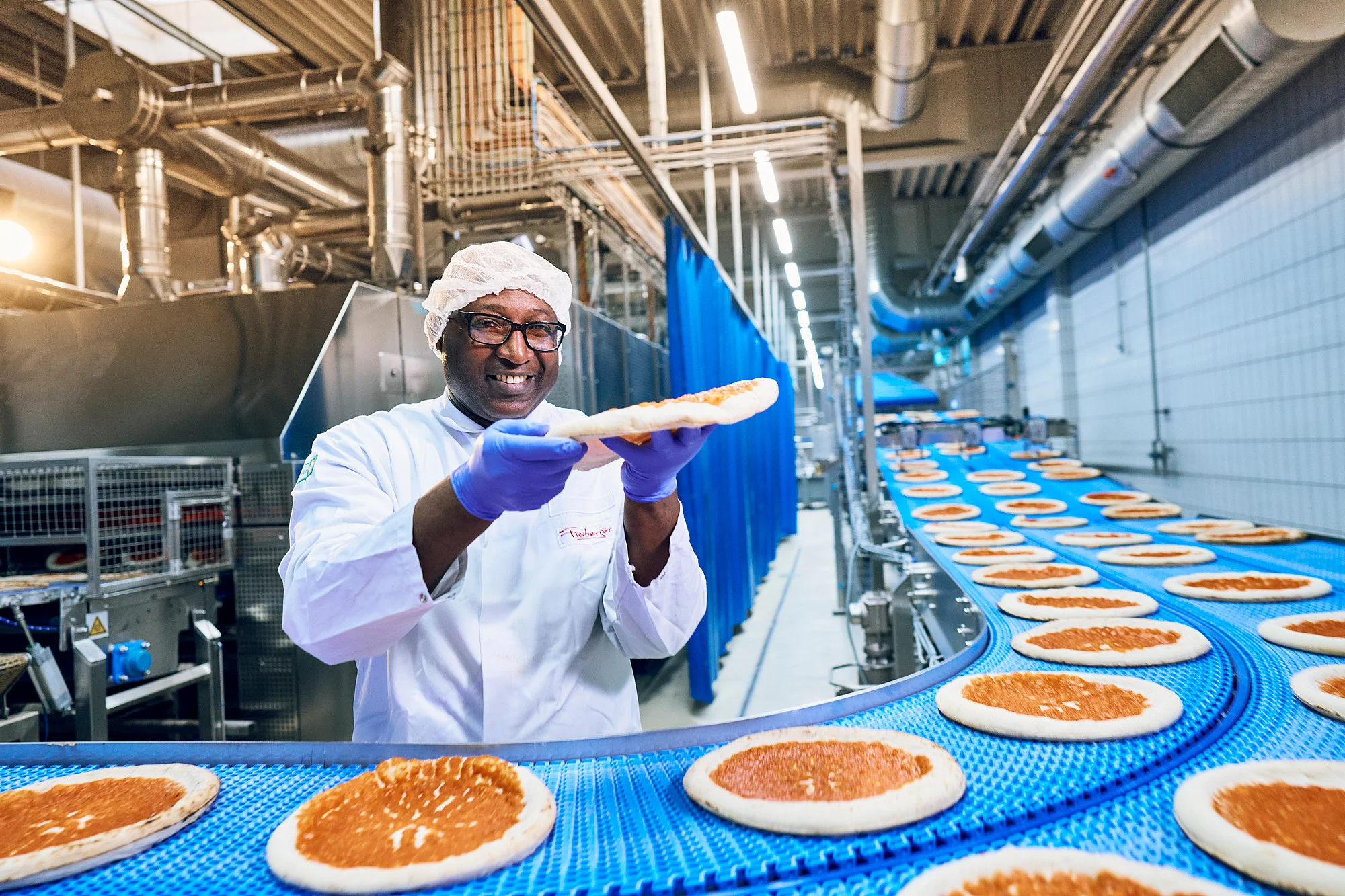 Ein Mitarbeiter in Arbeitskleidung in der Produktion am Fließband mit einer Pizza in der Hand.