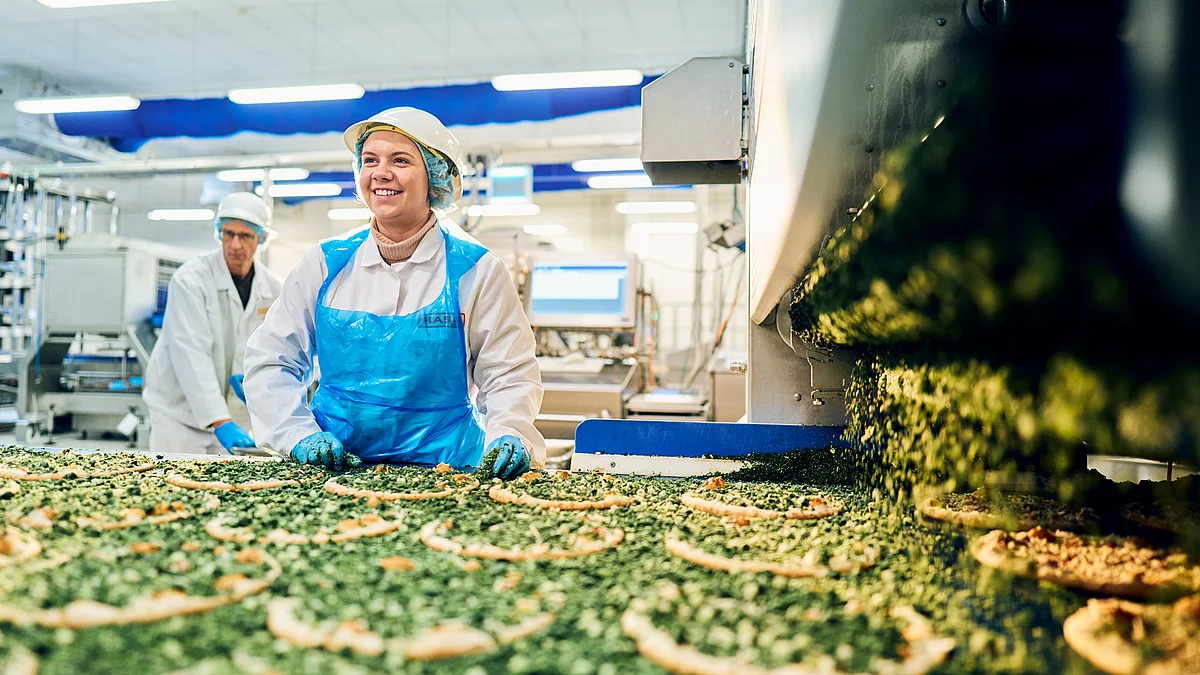 Zwei Mitarbeitende in Arbeitskleidung am pizzabefüllten Band. 