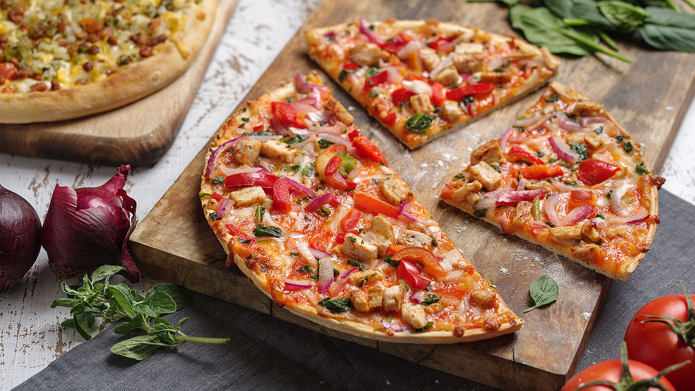 Zwei Pizzen auf jeweils einem Holzbrett, im Fokus die angeschnittene. Rundherum Tomaten, Basilikum, Zwiebeln. 