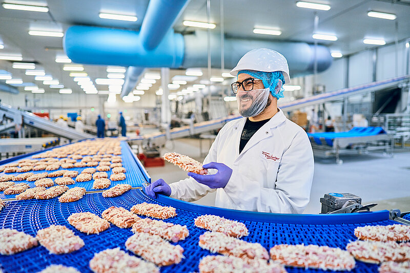 Ein Mitarbeiter in der Produktion am Fließband mit einem Pizza-Baguette in der Hand. 