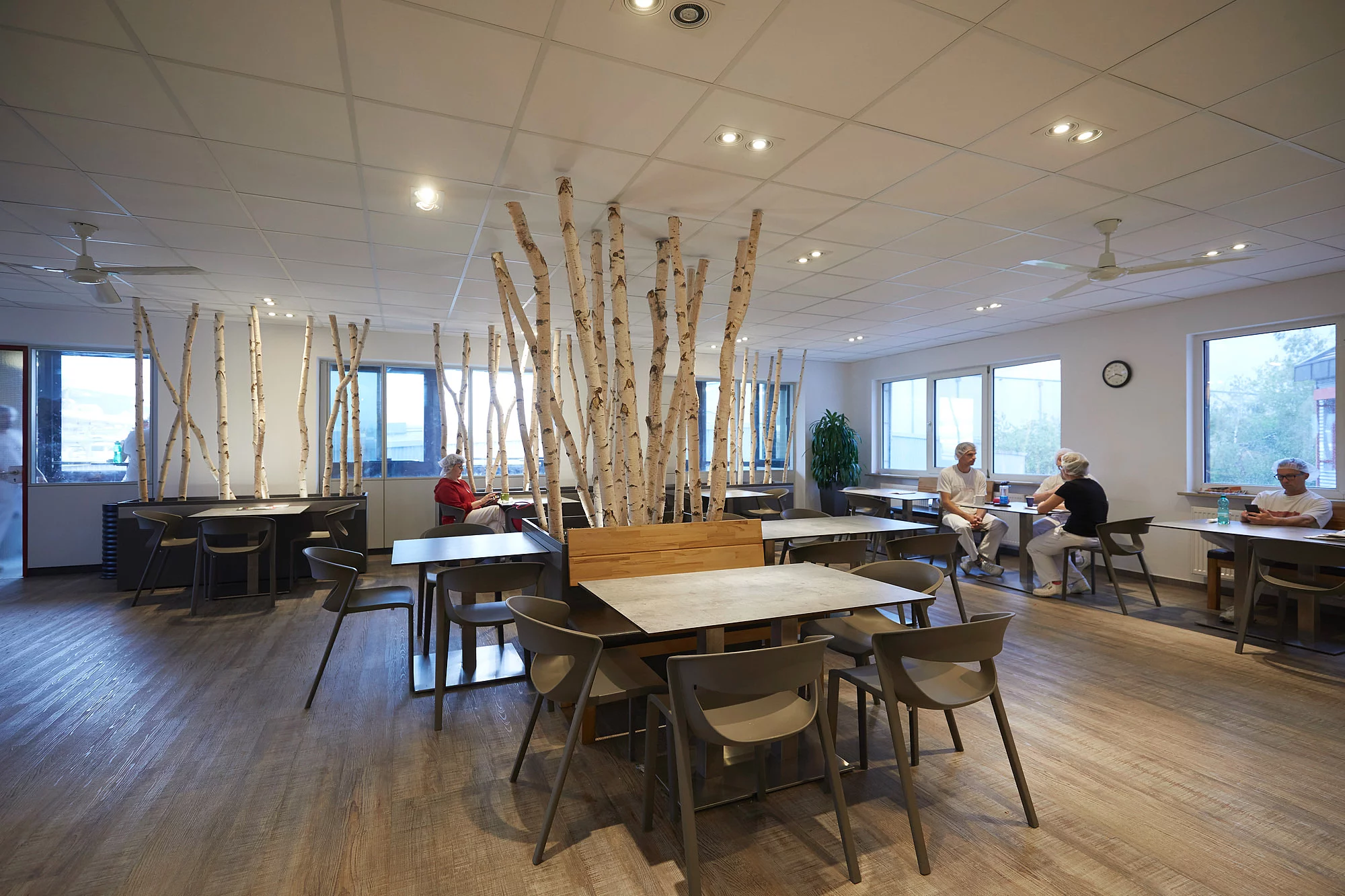 Mitarbeitende sitzen in Arbeitskleidung an unterschiedlichen Tischen in einem Aufenthaltsraum.