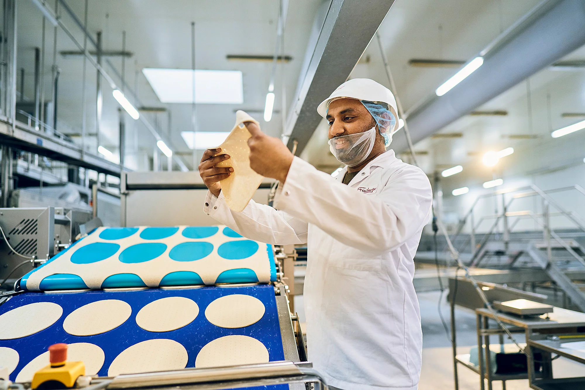 Mitarbeiter steht in Arbeitskleidung vor einem Fließband befüllt mit Pizzateig und schaut lächelnd auf einen Pizzateig in seiner Hand.