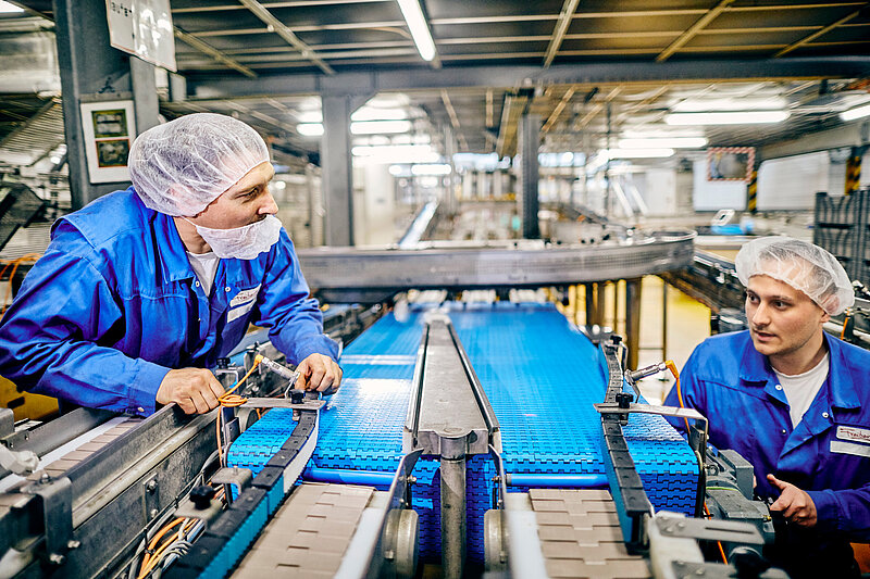Zwei Mitarbeiter in Arbeitskleidung am Schrauben eines Fließbandes.