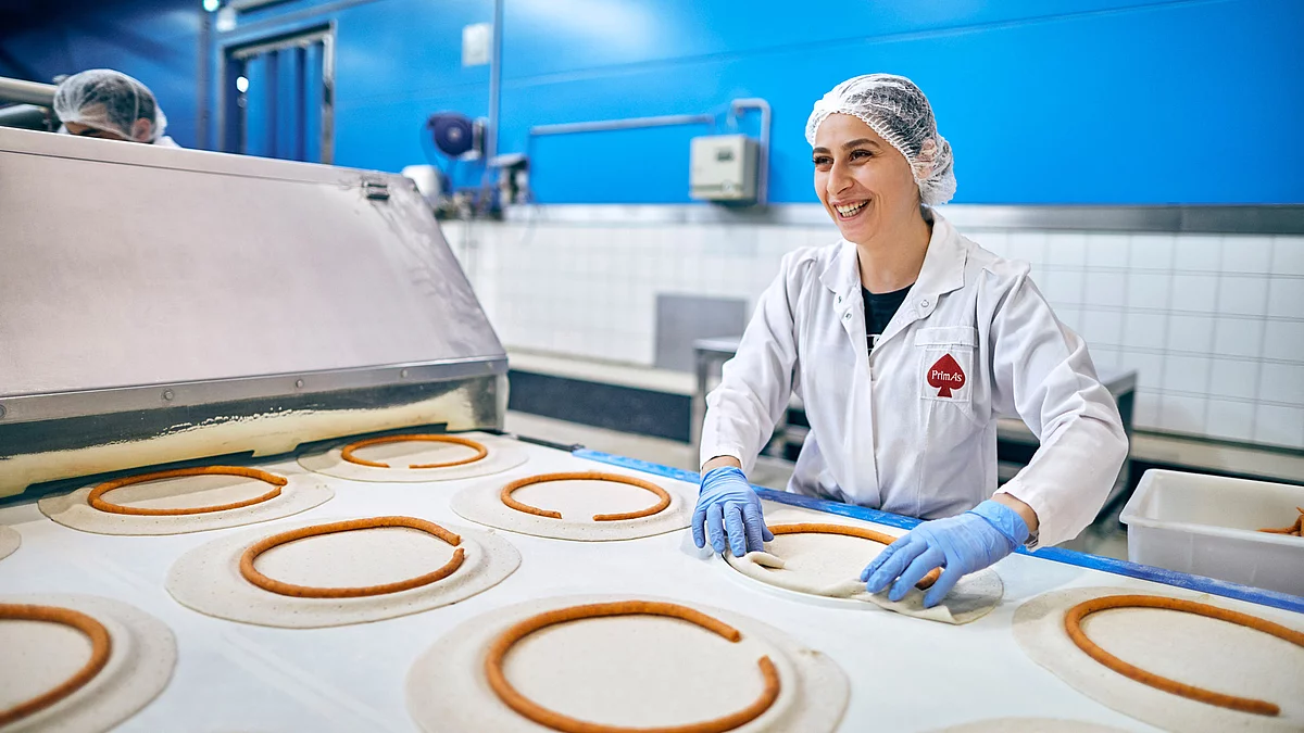 Mitarbeiterin in Arbeitskleidung steht vor dem Produktionsband für Pizzaböden.