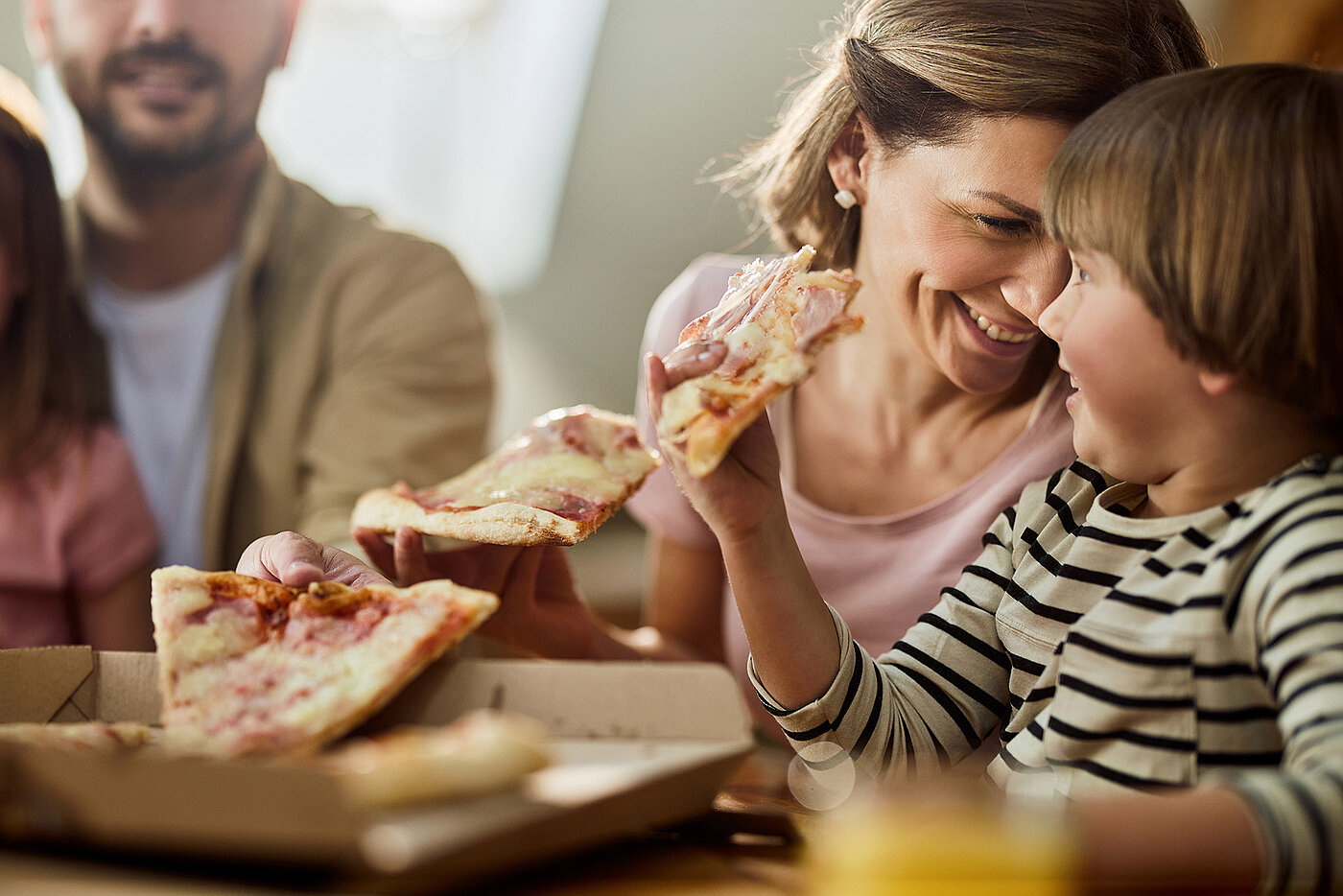 Eine Familie sitzt mit einer Pizza am Tisch. Die Mutter schaut mit einem Pizzastück in der Hand lächelnd zu ihrem Sohn.