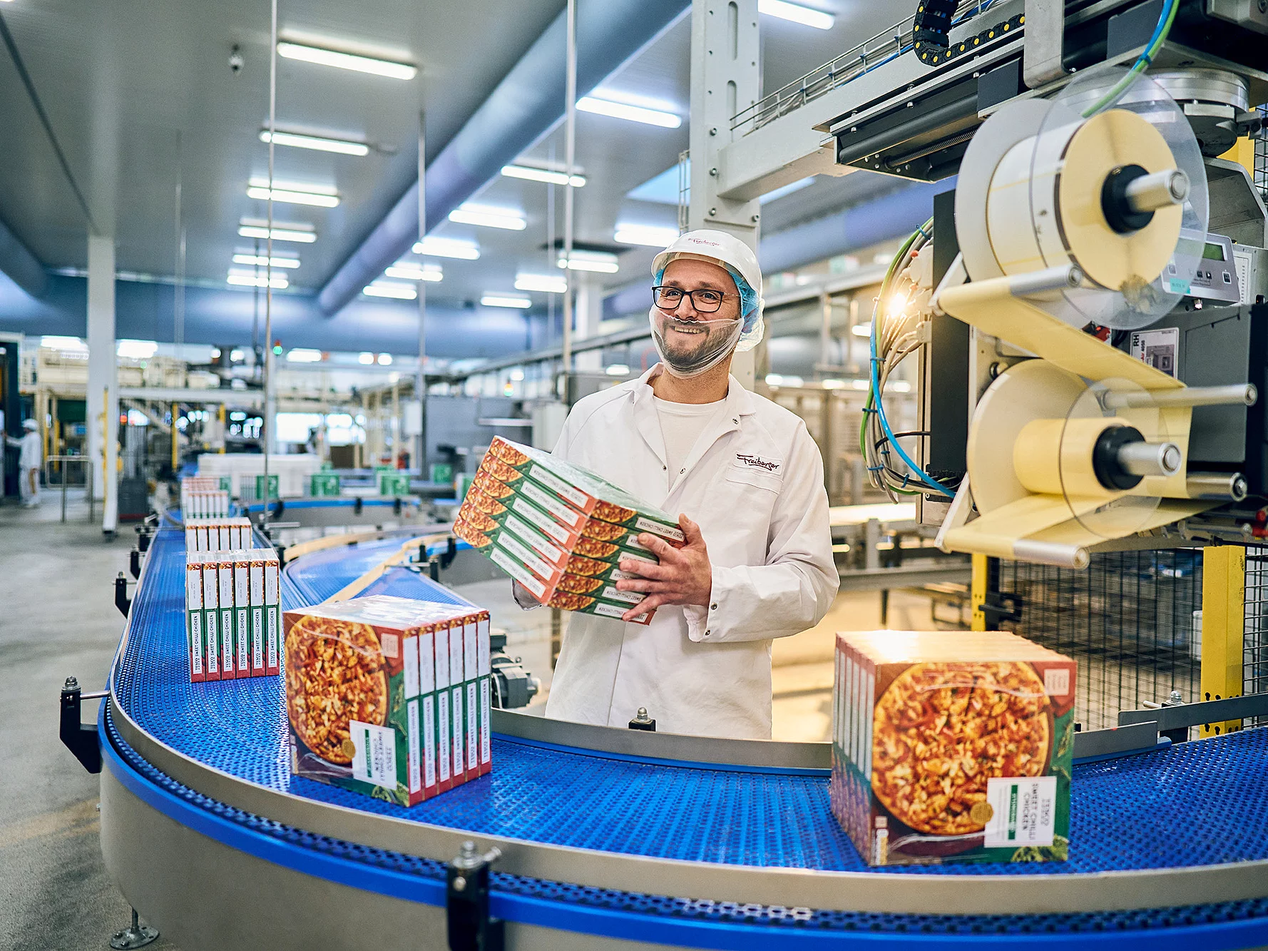 Mitarbeiter in Arbeitskleidung am Produktionsband mit vier Pizzakartons in der Hand. 