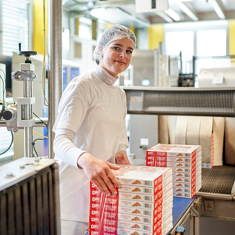 Eine Mitarbeiterin steht in der Produktion vor vielen gestapelten Pizzakartons.
