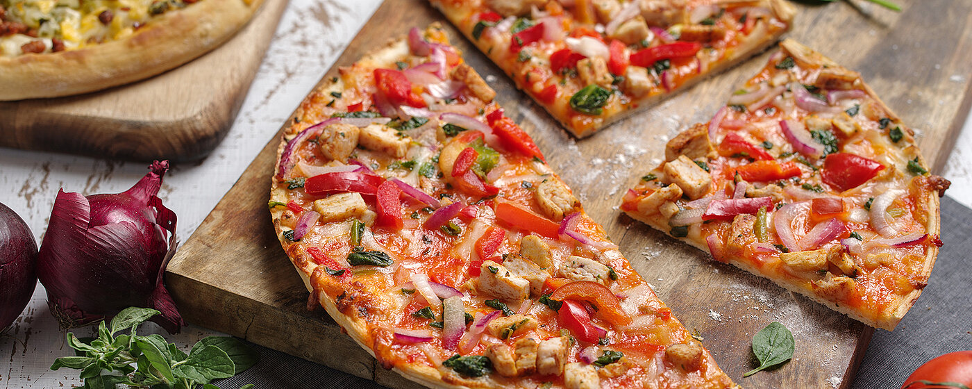 Zwei Pizzen auf jeweils einem Holzbrett. Im Fokus eine angeschnittene, drumherum Tomaten, Basilikum und Zwiebeln. 