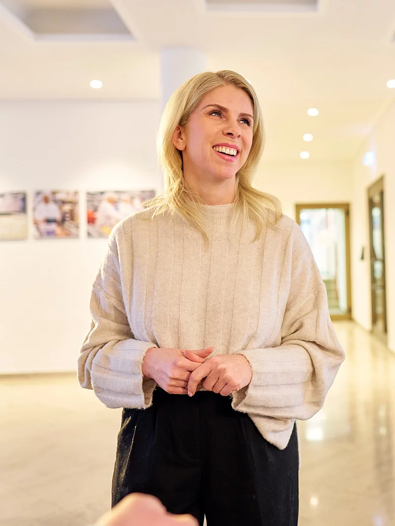 Eine Mitarbeiterin steht lächelnd in einer Lobby. Im Hintergrund sind weitere Personen zu sehen.