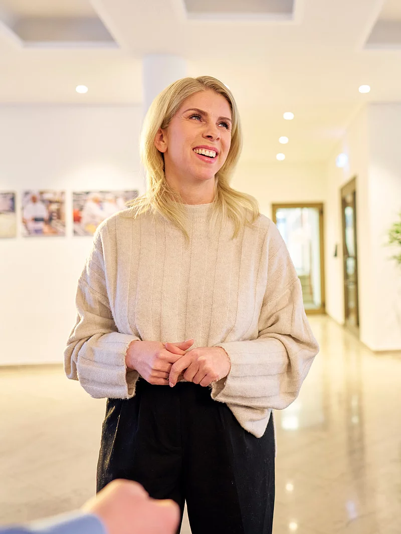 Eine Mitarbeiterin steht lächelnd in einer Lobby. Im Hintergrund sind weitere Personen zu sehen.