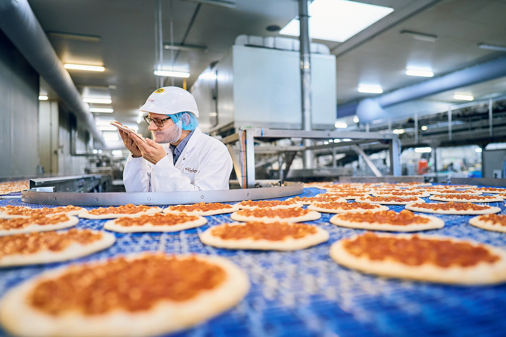 Mitarbeiter steht in Arbeitskleidung vor einem Fließband befüllt mit Pizzaböden. Er richt an einem Pizzaboden in seiner Hand.