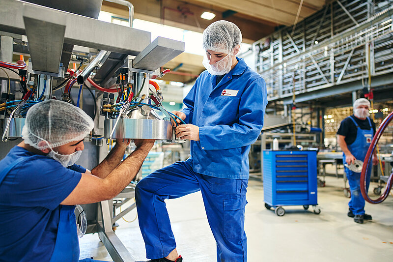 Zwei Mitarbeiter in Arbeitskleidung schrauben zusammen an einer Maschine herum. Im Hintergrund läuft ein weiterer Mitarbeiter. 