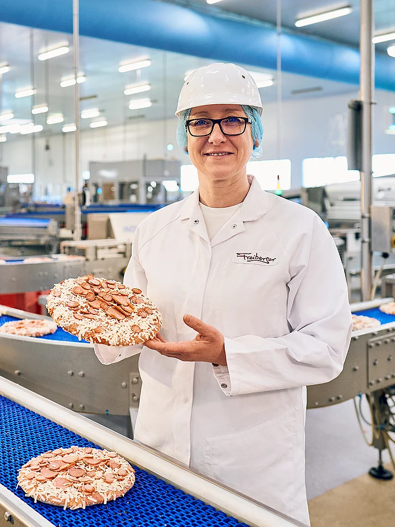 Eine Mitarbeiterin steht in Arbeitskleidung an einem Fließband befüllt mit Pizzen. Im Hintergrund steht ein weiterer Mitarbeiter.