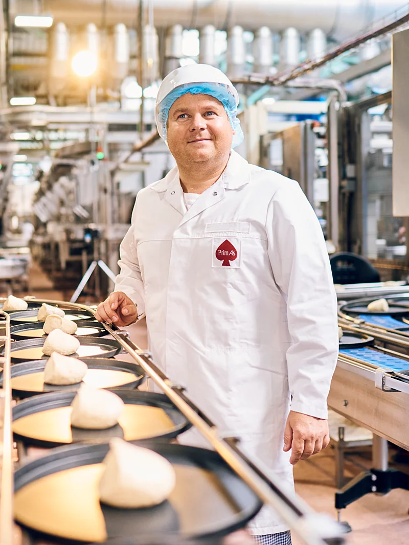 Ein Mitarbeiter steht in Arbeitskleidung an einem Fließband befüllt mit Pizzaböden.