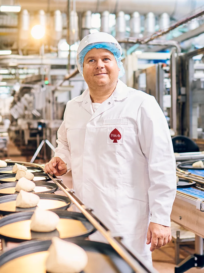 Ein Mitarbeiter steht in Arbeitskleidung an einem Fließband befüllt mit Pizzaböden.