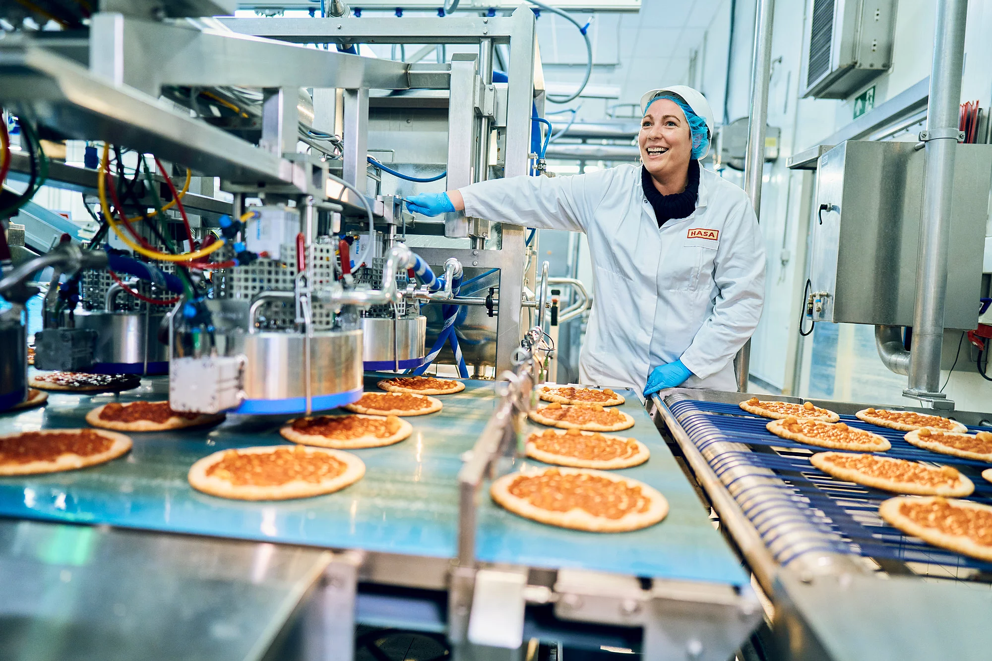 Mitarbeiterin in Arbeitskleidung in der Produktion vor einem Fließband mit Pizzen.