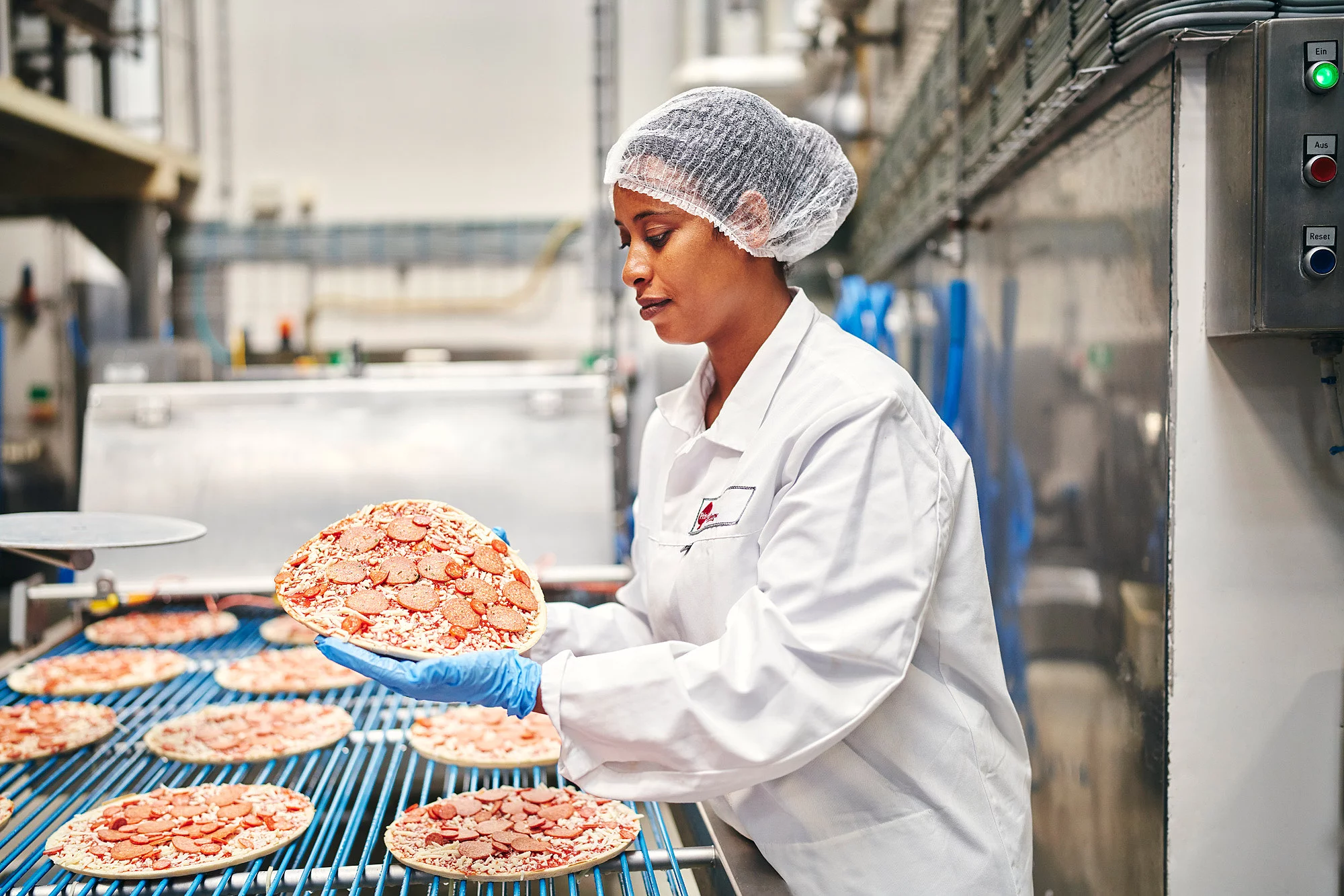 Mitarbeiterin in Arbeitskleidung an der Produktionsmaschine mit einer Pizza in der Hand.