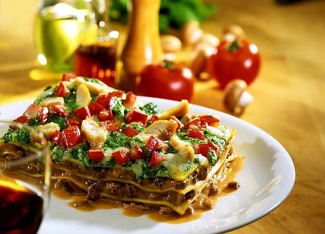 Lasagne auf einem Teller angerichtet dahinter mit Tomaten, Knoblauch und Pfeffermühle dekoriert.